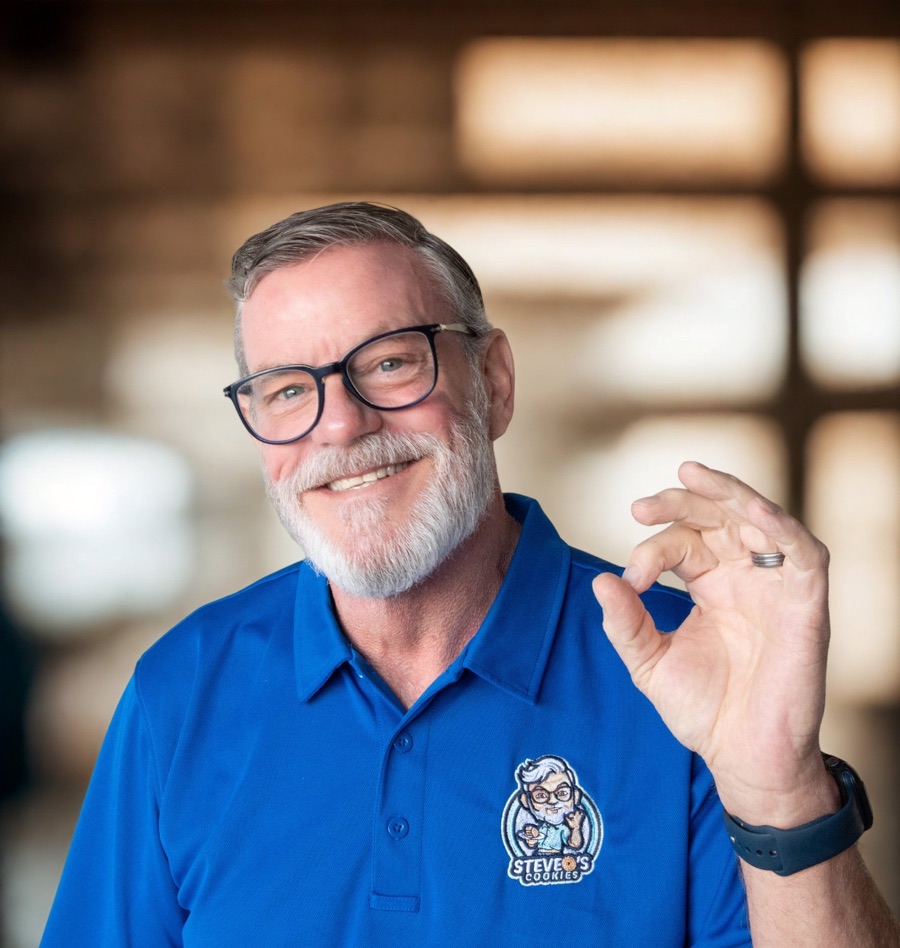 Steve, smiling in his blue steveO's cOOkies polo, making the chef-kiss gesture from his own logo